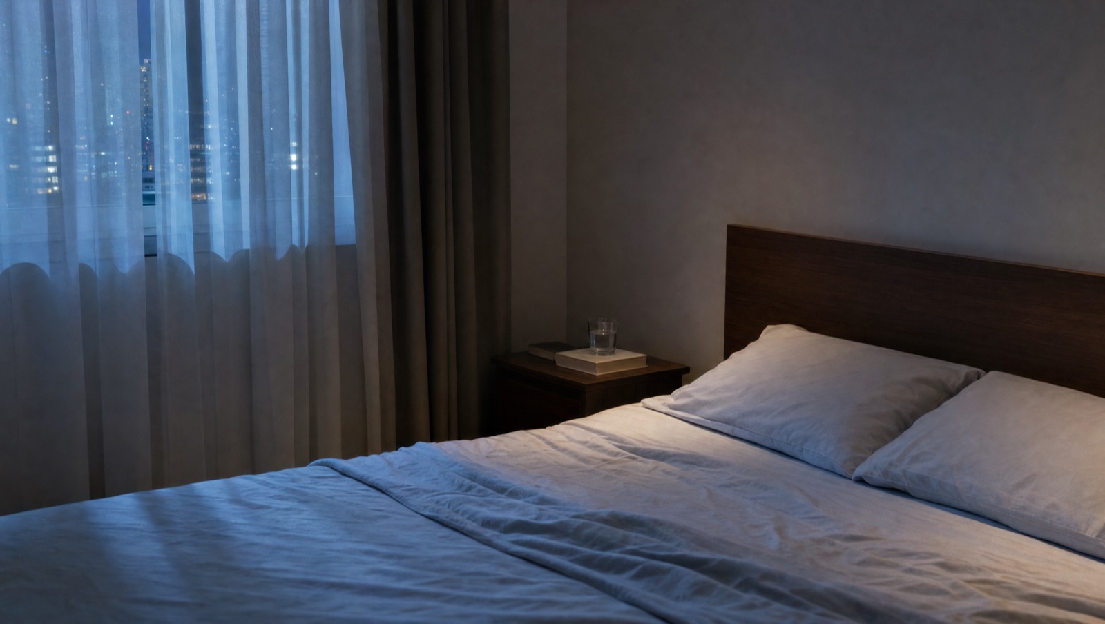 A dimly lit bedroom at night, with an empty bed, two pillows and city lights behind sheer curtains